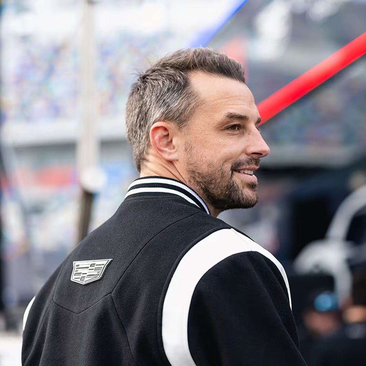 A man wearing a black-and-white jacket with Cadillac racing logos stands in front of equipment at a motorsports event.
