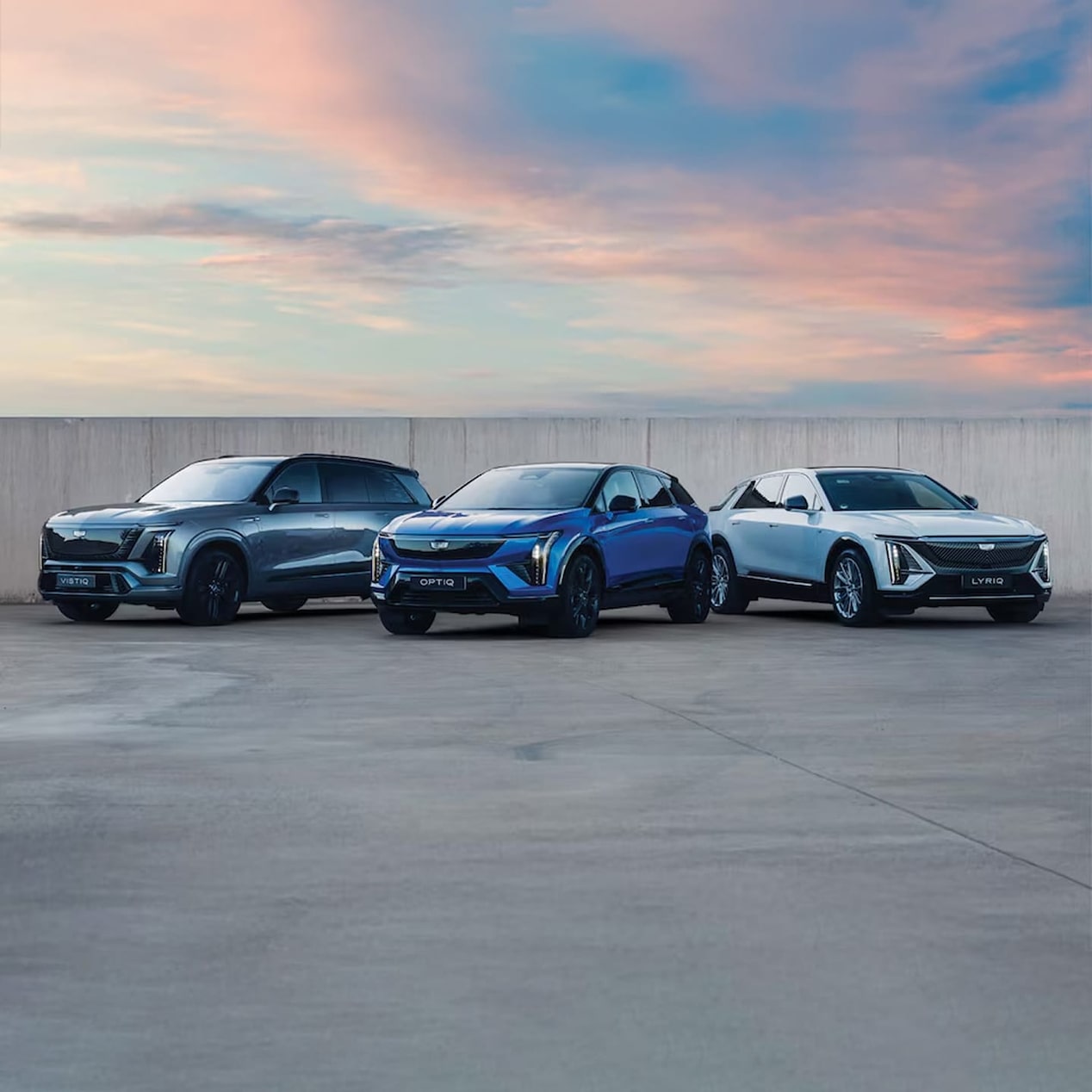 Cadillac’s all-electric luxury SUVs – OPTIQ, LYRIQ and VISTIQ – grouped on a clean, grey slate-like surface, with pink-barred sky.