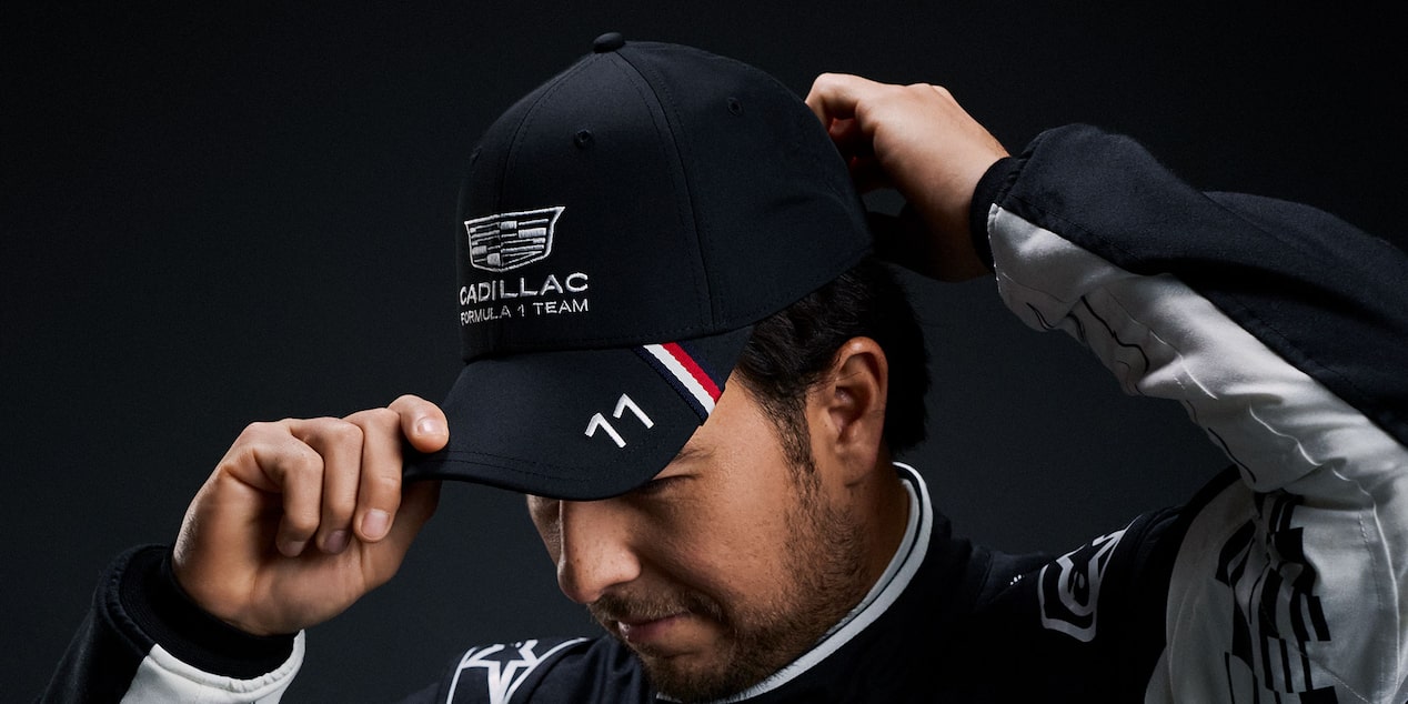 A team member wearing a black, Cadillac-branded cap adjusts the brim against a dark studio background.