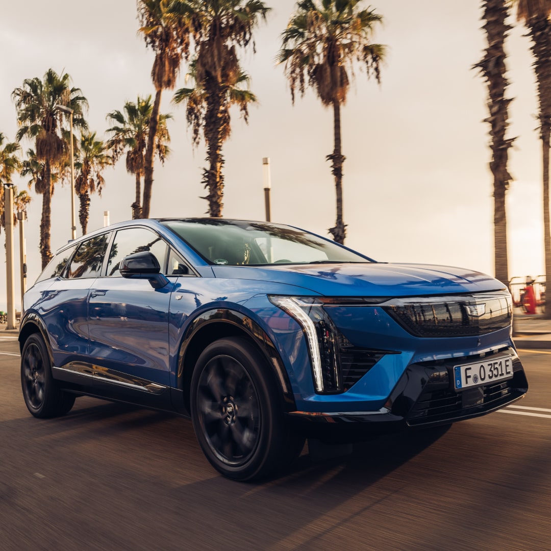 Vue de trois quarts avant d’un Cadillac OPTIQ bleu, un SUV électrique sportif, circulant le long d’une route bordée de palmiers en plein jour.