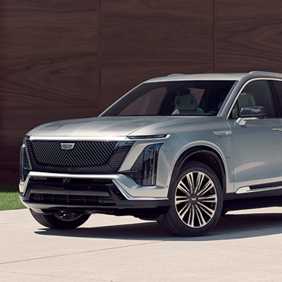 Vue avant trois-quarts de la Cadillac VISTIQ, SUV électrique 7 places, stationné devant un bâtiment moderne à l’architecture contemporaine avec façade en panneaux de bois.