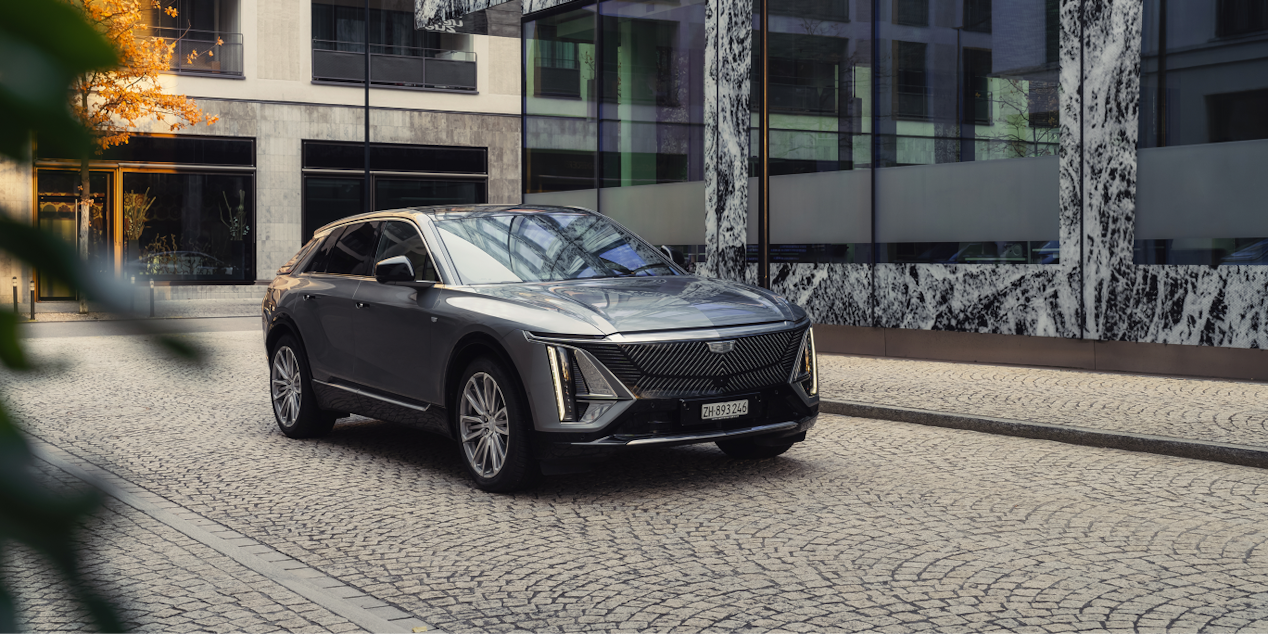 Vue avant trois-quarts d’un SUV électrique Cadillac stationné dans une rue pavée devant un bâtiment urbain moderne, illustrant les nombreuses possibilités de personnalisation de votre voiture électrique Cadillac.