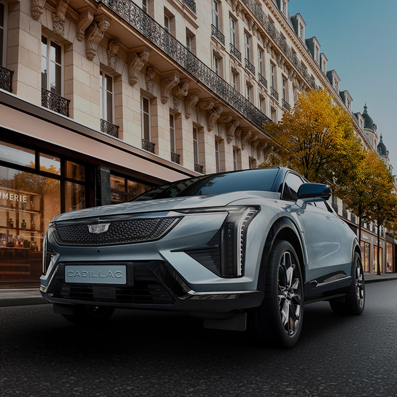 Cadillac OPTIQ STUDIO Edition – das markante Elektro-SUV fährt in einer europäischen Stadt an einem Parfümladen vorbei.