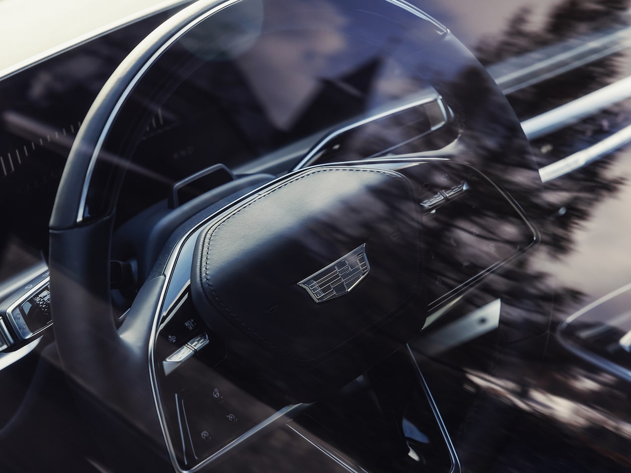 Volant d’un SUV de luxe 100 % électrique Cadillac, vu depuis la vitre latérale, où se dessinent de légers reflets d’arbres.