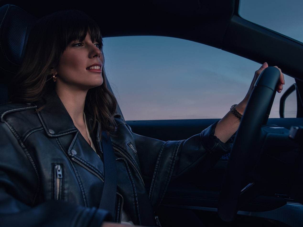 Vue de profil d’une femme en blouson biker en cuir noir, au volant d’un SUV de luxe Cadillac. À travers sa vitre latérale, un ciel aux reflets rosés évoque le coucher du soleil.