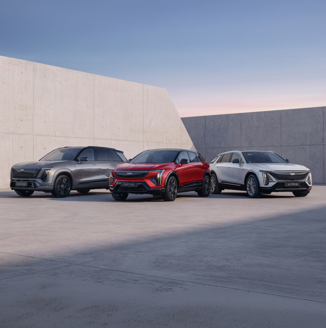 Three-quarter view of Cadillac OPTIQ, LYRIQ and VISITIQ, parked tail-to-tail in a fan formation on a concrete forecourt.