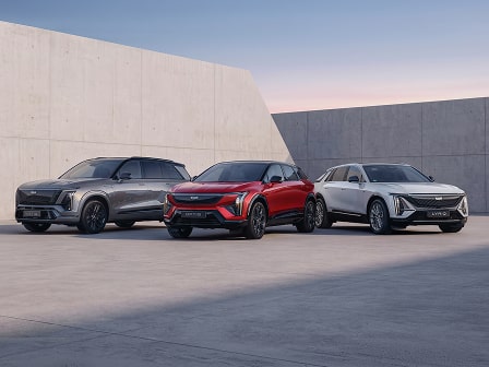 OPTIQ, LYRIQ und VISTIQ vereint. Cadillacs vollelektrische Luxus-SUVs auf einer eleganten schiefergrauen Fläche vor einem Himmel in sanften Rosatönen.