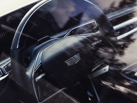 Ein Blick ins Cockpit: das Lenkrad eines vollelektrischen Cadillac Luxus-SUVs, eingefangen durch die Seitenscheibe mit sanften Spiegelungen der umliegenden Natur.
